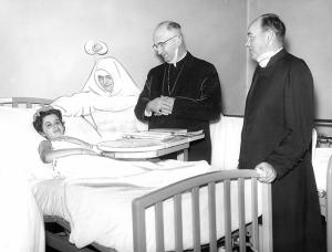 Archbishop Meyer visits Lucy Mardine, survivor of Our Lady of the Angels fire. With him are Sister H. Alumnda, P.H. J.C., administrator of St. Anne hospital, and Father Richard J. Birmingham, hospital chaplain. The Dec.1, 1958, fire at Our Lady of the Angels School, Chicago, killed 92 students and three BVM sisters, all teachers in the school. It led to changes in fire codes for schools and other public buildings and permanently changed those affected by it, its neighborhood and the city of Chicago. (Chicago Catholic file photo)