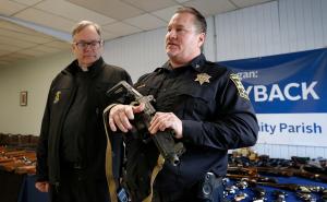 El padre Timothy O'Malley, pastor de Most Blessed Trinity mira mientras el subdirector de policía de Waukegan,  Chief Scott Chastain sostiene un arma automática entregada.