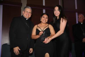 Yareli Godínez y Natalia Ramírez, coordinadoras asociadas de Iskali en Queen of All Nations, sonríen junto al obispo Casey mientras muestran el galardón de la Noche de Gala.